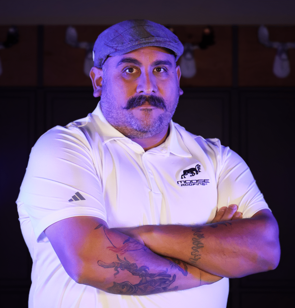Man in a cap and white shirt with "Moose Roofing" logo crossing his arms.