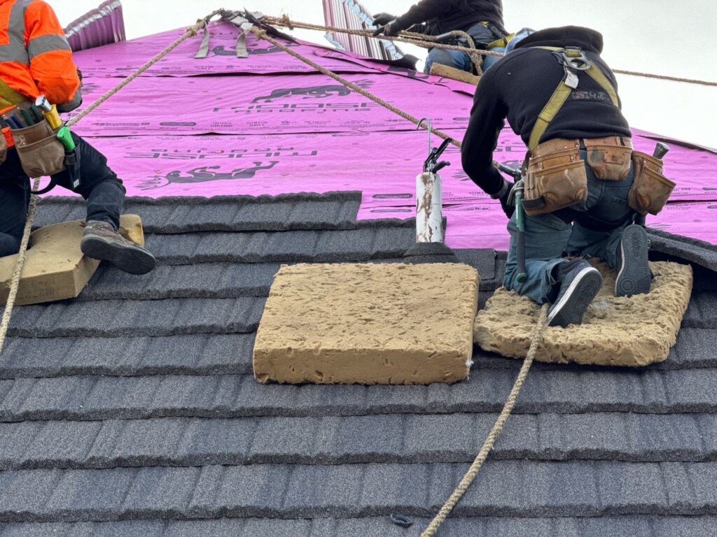 Roofers installing shingles on a steep roof with safety harnesses in Omaha.
