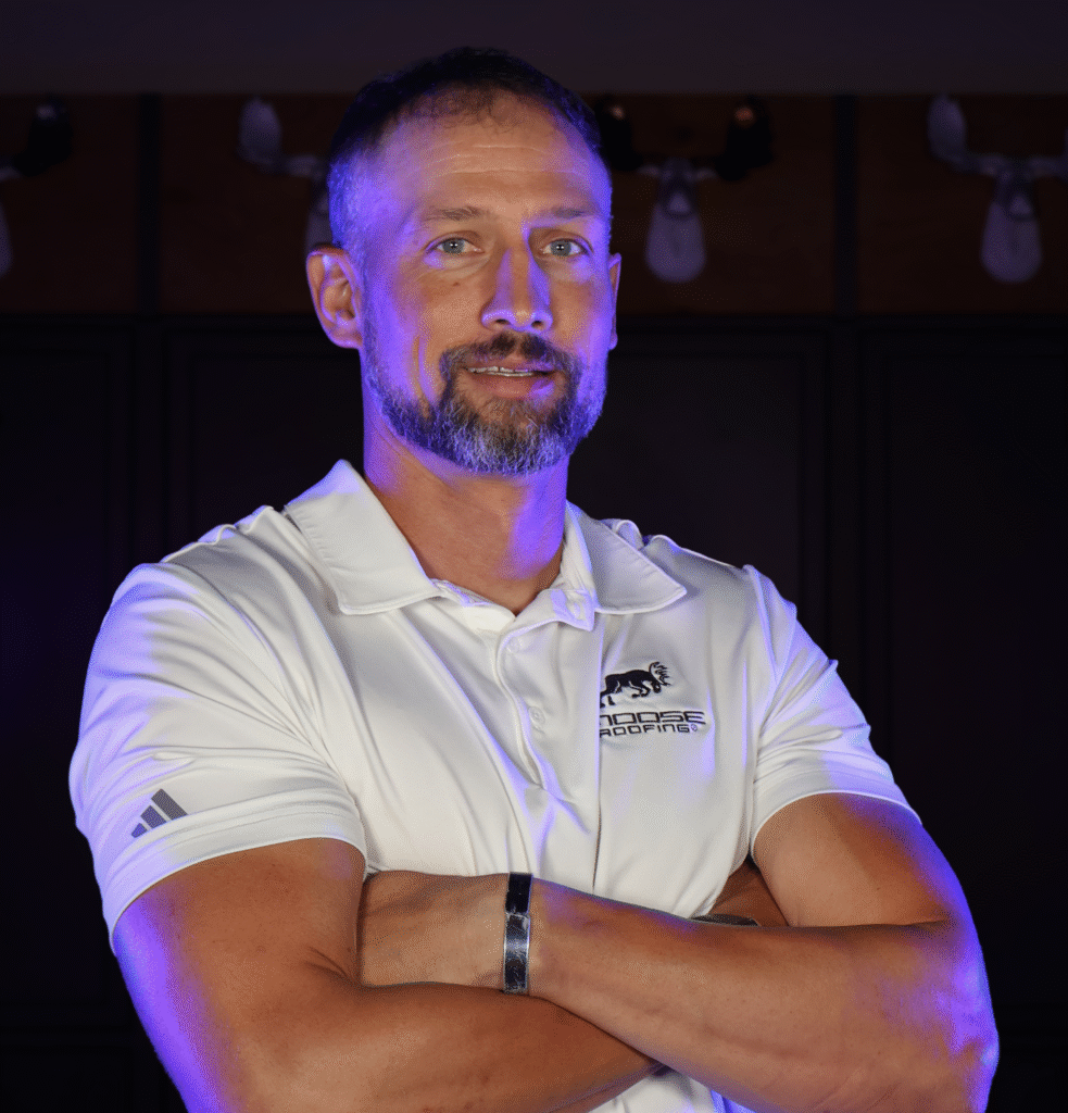 Person in a white polo with roofing logo stands confidently with arms crossed.