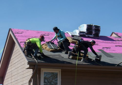 Roofers work on a house, installing shingles under a clear blue sky.