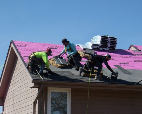Aaron Approved 2 Roofers work on a house, installing shingles under a clear blue sky.