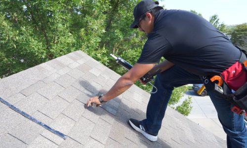 Roofer inspects shingles on a residential roof surrounded by trees.