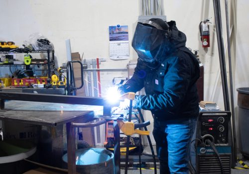 Worker welds metal in a workshop filled with tools and equipment.
