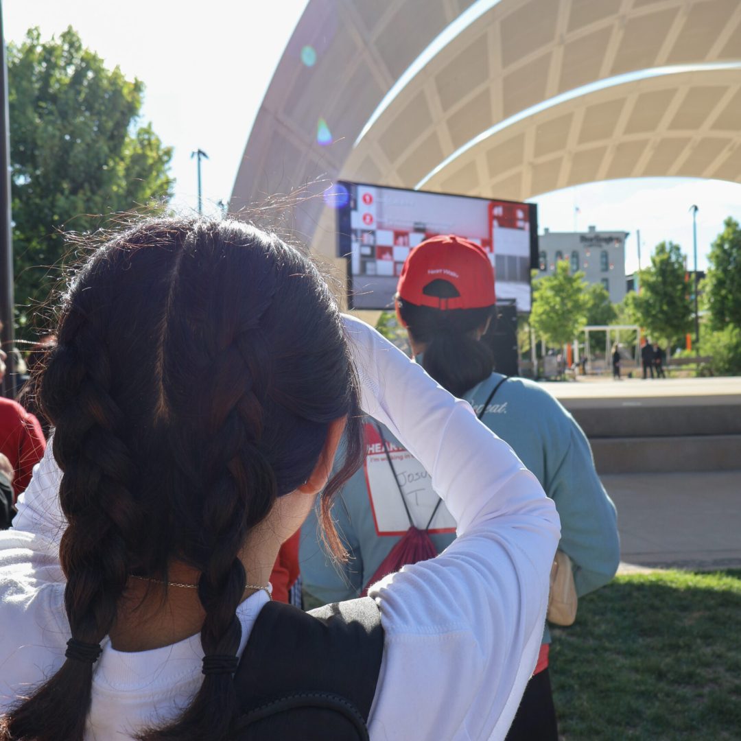 People gathered under a large, curved structure outdoors in sunny weather.