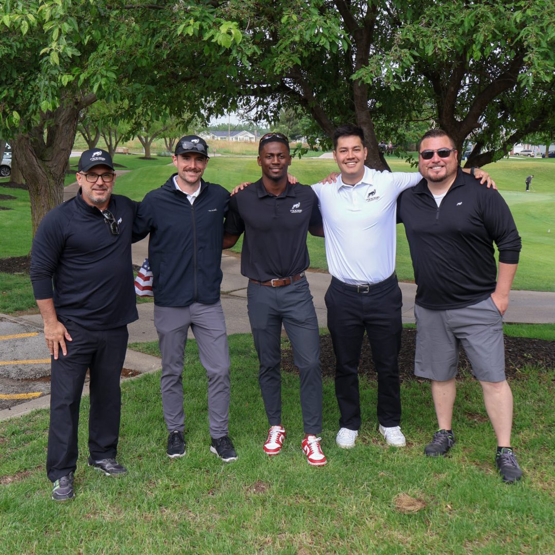 Five men in casual attire pose together on a grassy area under trees.
