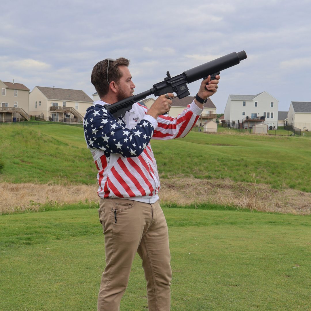 Man in American flag jacket aiming a large gun with houses in the background.
